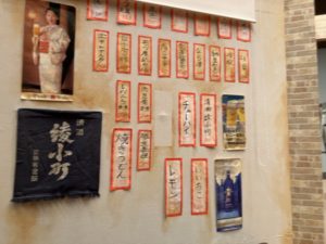 居酒屋門出の壁に貼られたメニュー。 ビールのポスターや、 清酒「綾小町」の前掛けなども飾られています。