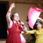 歌って踊る吉野、ピンク色の大きな扇を持って踊る山本